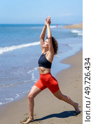Full length of fit barefoot female with curly hair in sportswear doing Crescent Lunge pose while practicing yoga near waving sea. Sportswoman practicing yoga on beach. Стоковое фото, агентство Ingram Publishing / Фотобанк Лори