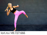 Flexible young blonde hair with long ponytail wearing activewear and doing martial arts exercise while training against gray background in modern fitness club. Flexible woman doing doing martial arts exercise. Стоковое фото, агентство Ingram Publishing / Фотобанк Лори