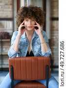 Serious young female with curly hair in denim jacket and jeans sitting on leather chair in room against brick wall while touching face. Woman in denim jacket and bra sitting on chair and touching face. Стоковое фото, агентство Ingram Publishing / Фотобанк Лори