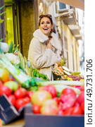 Low angle of cheerful young woman in warm clothes, smiling and looking at camera while standing near stall with bunch of fresh ripe fruits and vegetables in daylight. Gleeful young woman in cozy coat standing by fruits in shop. Стоковое фото, агентство Ingram Publishing / Фотобанк Лори