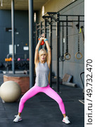 Fit young female doing squat exercise with kettlebell during fitness training in modern gym and stretching body against blurred interior of equipment on gray background. Fit young woman doing squats exercises with kettlebell in gym. Стоковое фото, агентство Ingram Publishing / Фотобанк Лори