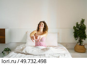Smiling adult female with curly hair and eyes closed while sitting on cozy bed in daylight and stretching reverse clasped hands, in front with interlocked fingers near green plant. Cheerful woman sitting on bed in sleepwear. Стоковое фото, агентство Ingram Publishing / Фотобанк Лори