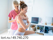Concentrated osteopath in pink uniform massaging back of young female client in inner wear sitting with arms folded on bed during physiotherapy session in clinic. Focused osteopath massaging back of female client sitting on bed in clinic. Стоковое фото, агентство Ingram Publishing / Фотобанк Лори