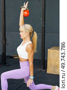 Side view of fit strong female doing kneeling kettlebell press exercise during fitness workout in modern gym against gray wall on floor. Flexible woman doing kneeling kettlebell press exercise. Стоковое фото, агентство Ingram Publishing / Фотобанк Лори
