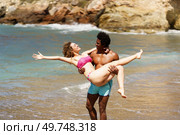 Side view of cheerful young female stretching arm backwards, while lying in hands of standing African American male and looking at each other near seawater during daytime. Diverse couple having fun on beach in daylight. Стоковое фото, агентство Ingram Publishing / Фотобанк Лори