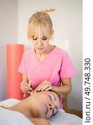 Chiropractor professional beautician doing deep ear massage of young female customer with ear probe pen during skin care treatment in modern beauty salon. Professional beautician applying ear probe on female client in salon during auriculotherapy. Стоковое фото, агентство Ingram Publishing / Фотобанк Лори