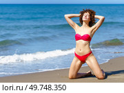 Calm slim female with curly hair in bikini standing on knees and hands behind head while getting suntan on coast. Relaxing woman sunbathing near waving sea. Стоковое фото, агентство Ingram Publishing / Фотобанк Лори