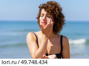 Young curly haired female in black bikini applying sunscreen cream on cheeks while standing near sea on sunny summer day protecting skin from sunburn. Young woman smearing sunblock cream on face. Стоковое фото, агентство Ingram Publishing / Фотобанк Лори