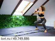 Full body of young curly haired female athlete in sportswear doing agility ladder squat jumps in gym with pitched roof. Smiling woman squatting in gym during training with agility ladder. Стоковое фото, агентство Ingram Publishing / Фотобанк Лори