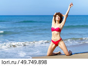 Full body of tender woman with curly hair and in red bikini raising arm while standing on knees on sandy beach of ocean and sunbathing. Sensual woman resting on sandy shoreline. Стоковое фото, агентство Ingram Publishing / Фотобанк Лори