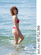 Side view of self assured young happy female, with brown curly hair in red bikini smiling brightly and looking at camera while walking in sea in sunlight. Cheerful woman in swimwear smiling while walking in sea. Стоковое фото, агентство Ingram Publishing / Фотобанк Лори