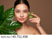 Close up woman face with green exotic leaf. Model doing skin care routine, scrapping, massage facial skin with jade gua sha scrapper. Стоковое фото, агентство Ingram Publishing / Фотобанк Лори