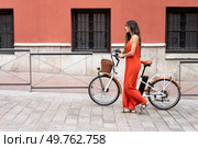 A fashionable woman in a vibrant red dress strolls beside her chic ebike along a colorful wall, embodying an elegant urban lifestyle that showcases her unique style and charisma. A Stylish Woman in a Vibrant Red Dress Walks by Her ebike Near a Colorful Artistic Wall. Стоковое фото, агентство Ingram Publishing / Фотобанк Лори