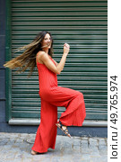 A stylish and confident woman strikes a playful pose in a vibrant red jumpsuit that perfectly showcases her flair and joy, all against a bright green backdrop that adds to the lively scene. A woman in a bright red jumpsuit poses playfully outdoors, radiating joy and confidence. Стоковое фото, агентство Ingram Publishing / Фотобанк Лори