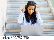 A young woman with curly hair shows frustration while using her laptop on the stairs, battling tech issues and concentration problems, leading to emotional distress and increased stress. A young woman feeling stressed and anxious as she struggles with frustrating laptop issues. Стоковое фото, агентство Ingram Publishing / Фотобанк Лори