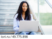 A young woman sits comfortably on the outdoor steps, actively typing on her laptop. She conveys a truly relaxed vibe that captures the essence of modern remote work amidst a vibrant, lively setting. A woman is working on her laptop while comfortably sitting on outdoor steps in sunlight. Стоковое фото, агентство Ingram Publishing / Фотобанк Лори