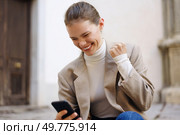 A young woman, elegantly dressed in a fashionable blazer and cozy turtleneck, beams with joy as she experiences a delightful moment while intently gazing at her smartphone, capturing her happiness. Joyful Moment A Young Woman Celebrating with Her Smartphone in a Cheerful Atmosphere. Стоковое фото, агентство Ingram Publishing / Фотобанк Лори