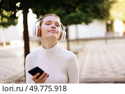 A young woman, dressed in a cozy white sweater and wearing headphones, smiles serenely while enjoying music outdoors, holding her smartphone in a bright and sunlit environment filled with nature. A Young Woman Happily Enjoying Music Outdoors while Wearing Headphones and Using Her Smartphone. Стоковое фото, агентство Ingram Publishing / Фотобанк Лори