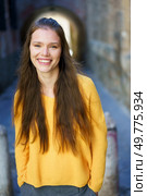 A joyful young woman with long, flowing hair, dressed in a vibrant and bright yellow sweater, stands in a picturesque urban alley, radiating positivity, warmth, and happiness like sunshine. A cheerful woman in a yellow sweater posed in an urban setting with vibrant city colors. Стоковое фото, агентство Ingram Publishing / Фотобанк Лори
