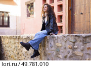 In a vibrant urban setting, a young woman with curly hair in a stylish leather jacket and trendy jeans enjoys a moment on a stone wall, soaking in the lively atmosphere around her. A Young Woman is Comfortably Sitting and Relaxing on a Stone Wall in a Lovely Urban Setting. Стоковое фото, агентство Ingram Publishing / Фотобанк Лори
