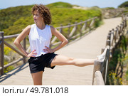 Focused young female with hands on waist in sportswear, looking away while standing with stretched leg on bridge fence and hearing music from earbuds in sunlight against blurred nature. Slim young woman stretching leg on bridge in sunny day. Стоковое фото, агентство Ingram Publishing / Фотобанк Лори