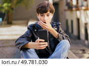 A young person displaying a shocked expression intently checks their smartphone while seated on outdoor steps in a bustling urban environment, illustrating the dynamics of modern digital engagement. A surprised young individual engaging deeply with their smartphone while sitting on urban steps. Стоковое фото, агентство Ingram Publishing / Фотобанк Лори
