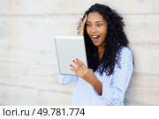 A young woman with beautifully curly hair radiates surprise and beams with joy while enthusiastically using a tablet, showcasing a lively moment filled with genuine engagement and deep emotion. An Excited Womans Reaction while Engaging with a Tablet up against a Concrete Wall. Стоковое фото, агентство Ingram Publishing / Фотобанк Лори