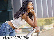 A thoughtful young woman with beautifully curly hair leans gracefully on a railing, gazing thoughtfully into the distance, surrounded by the hustle and bustle of an urban environment. A Contemplative Young Woman Posing Thoughtfully Against an Urban City Backdrop Scene. Стоковое фото, агентство Ingram Publishing / Фотобанк Лори