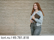 A stylish and fashionable young woman with beautiful curly red hair is smiling brightly while walking outdoors, holding a notebook, which radiates her confidence and charm effortlessly. A Fashionable Young Woman Walking Outside with a Notebook for Studying and Inspiration. Стоковое фото, агентство Ingram Publishing / Фотобанк Лори