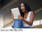 A joyful young woman is expressing her excitement while looking intently at a tablet device in her hands, showcasing her surprise and deep engagement, all within a lively and modern setting. An Excited Young Woman Happily Discovering Something New on Her Tablet Device Today. Стоковое фото, агентство Ingram Publishing / Фотобанк Лори