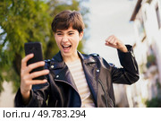 A young person in a stylish leather jacket happily takes a selfie outdoors, enjoying a vibrant atmosphere filled with energy and creating a cherished moment to remember and treasure. Young people celebrating outdoors, taking selfies, and embodying joy and friendship. Стоковое фото, агентство Ingram Publishing / Фотобанк Лори