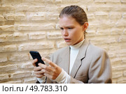 A young professional shows a troubled expression while concentrating on a smartphone, highlighting themes of modern technology, personal stress, and the anxiety it adds to their daily life. A concerned individual using a smartphone while standing against a textured wall outdoors. Стоковое фото, агентство Ingram Publishing / Фотобанк Лори