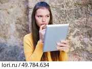 A young woman in a bright yellow sweater engages with her tablet while leaning against a rustic stone wall, blending contemporary tech with deep contemplation and personal reflection. A young woman is deeply engrossed in the tablet screen while sitting against a stone wall. Стоковое фото, агентство Ingram Publishing / Фотобанк Лори