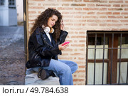 A fashionable young woman with curly hair sits on a stone wall, engrossed in her smartphone, reflecting modern youth culture in a vibrant urban setting filled with creativity and energy. A Young Woman with Beautiful Curly Hair is Using a Smartphone While in an Urban Setting. Стоковое фото, агентство Ingram Publishing / Фотобанк Лори