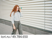 A vibrant young woman with long red hair walks outdoors, joyfully holding her smartphone. Dressed in a stylish outfit, she exudes confidence and happiness amid impressive modern architecture. A Young Woman Enjoys a Fun Phone Call Outside in a Stylish Outfit Showcasing Her Fashion Sense. Стоковое фото, агентство Ingram Publishing / Фотобанк Лори