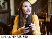 In a cozy cafe, a cheerful young woman in a yellow sweater happily uses her smartphone, symbolizing modern connections with family and friends despite todays fastpaced challenges. A Cheerful Young Woman is Engaged with Her Smartphone While Relaxing in a Cozy Cafe Setting. Стоковое фото, агентство Ingram Publishing / Фотобанк Лори