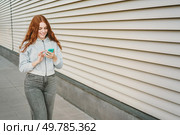 A young woman sporting long, beautiful reddish hair is delightfully enjoying her smartphone while casually walking outdoors in an urban environment filled with stunning contemporary architecture. A young woman casually walks and texts on her smartphone, enjoying modern architecture. Стоковое фото, агентство Ingram Publishing / Фотобанк Лори