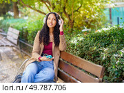 A young woman enjoys her day while sitting on a comfortable bench in a beautiful park. She is wearing stylish headphones, listening to her favorite music, and surrounded by lush, vibrant greenery. A Young Woman Delightfully Enjoying Music in a Relaxing Park Setting on a Pleasant Day. Стоковое фото, агентство Ingram Publishing / Фотобанк Лори