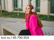 On a beautiful day, a vibrant young woman with long red hair enjoys music outdoors. Wearing stylish headphones and a bright pink jacket, she embodies carefree enjoyment and a casual lifestyle. A Young Woman Is Happily Enjoying Music While Wearing a Stylish Pink Jacket Outdoors. Стоковое фото, агентство Ingram Publishing / Фотобанк Лори