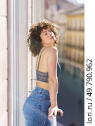 Calm young female in bra and jeans with Afro hairstyle standing on balcony with closed eyes while leaning on fence on sunny day. Dreamy woman leaning on balcony fence. Стоковое фото, агентство Ingram Publishing / Фотобанк Лори