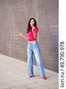 A young woman in a bright pink crop top and flared jeans dances joyfully against a textured wall, showcasing urban fashion and a vibrant lifestyle that inspires creativity and fun. A Joyful Woman is Dancing Enthusiastically in an Urban Setting, Showcasing a Trendy Outfit. Стоковое фото, агентство Ingram Publishing / Фотобанк Лори