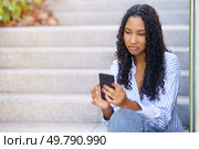 A young woman with beautifully curly hair is seated on outdoor steps, deeply engrossed in her smartphone, symbolizing the essence of modern casual lifestyle and contemporary connectivity. A young woman is actively using her smartphone while sitting comfortably on the steps outdoors. Стоковое фото, агентство Ingram Publishing / Фотобанк Лори