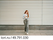 A stylish young woman with long red hair strolls by a colorful striped wall, checking her phone, embodying a vibrant, modern urban lifestyle full of connections and activities defining todays youth. A young woman is calmly walking in an urban setting while using her phone for connection. Стоковое фото, агентство Ingram Publishing / Фотобанк Лори