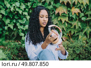 A young woman is peacefully examining and enjoying her stylish headphones, all while being surrounded by vibrant and lush greenery, which beautifully embodies a sense of tranquility and modern style. A young woman enjoys music with headphones, sitting peacefully amid vibrant green nature. Стоковое фото, агентство Ingram Publishing / Фотобанк Лори