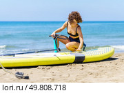 Smiling young female with curly ginger hair in swimwear sitting and tapping with hand on paddleboard, while holding handpump and looking down against blurred waving sea blue sky. Focused young woman sitting and checking paddleboard. Стоковое фото, агентство Ingram Publishing / Фотобанк Лори