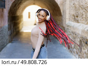In a lively urban setting, a stylish woman with vibrant red dreadlocks dances gracefully in her headphones, radiating joy, freedom, and confidence as she moves to the musics rhythm. A woman with striking red dreadlocks is enjoying music while standing in an urban archway. Стоковое фото, агентство Ingram Publishing / Фотобанк Лори