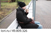 Pensive hipster drinking coffee sitting on a bench in the urban park. Handsome man wearing black coat eating street food, hot dog with paper cup in hand in the city. Stylish bearded middle-aged male. Стоковое видео, видеограф Марина Демидюк / Фотобанк Лори