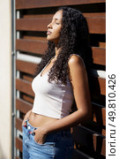 A young woman with beautiful, curly hair delights in the soothing warmth of the sun while leaning gracefully against a sturdy wooden panel, radiating a sense of tranquility and contemporary style. A serene woman is posing gracefully in the sunlight while standing against a wooden background. Стоковое фото, агентство Ingram Publishing / Фотобанк Лори