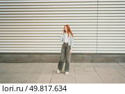 A fashionable young woman with vibrant red hair is standing confidently against a contemporary striped wall, actively engaging in a phone conversation while maintaining a relaxed and casual posture. A casual young woman, dressed in a trendy and stylish outfit, is engaging in a phone conversation. Стоковое фото, агентство Ingram Publishing / Фотобанк Лори