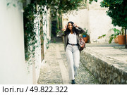 A fashionable young woman strides confidently down a picturesque alley, surrounded by lush greenery and charming architecture, enjoying every moment of this delightful urban exploration. A Stylish Young Woman is Enjoying a Leisurely Stroll Through a Quaint Cobblestone Street. Стоковое фото, агентство Ingram Publishing / Фотобанк Лори